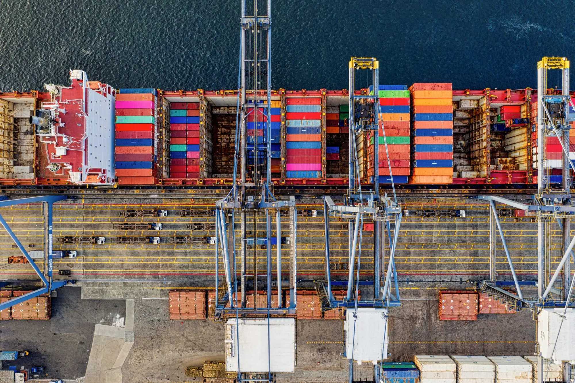 A container ship at a quay seen from directly above, stacked with coloured containers under a row of cranes