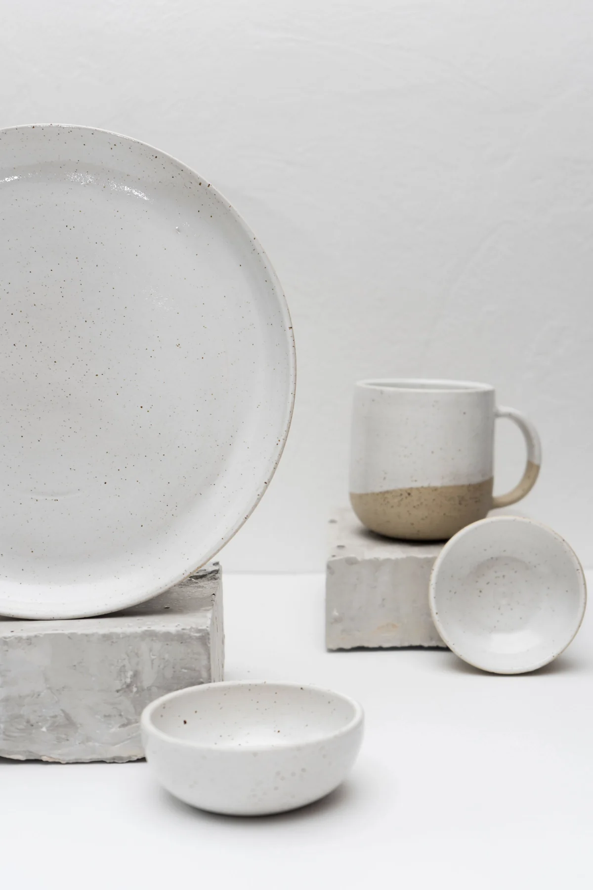 Three white ceramic pieces, a plate, a cup and a bowl, set apart on plain stone plinths