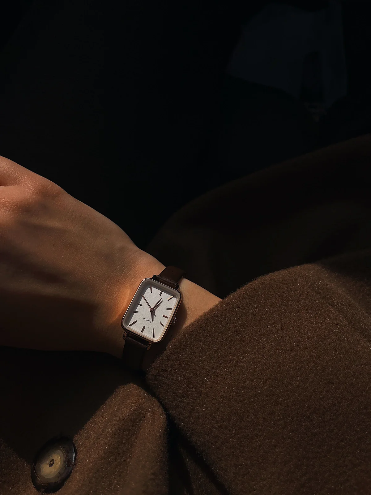 A small rectangular watch on a wrist against a dark wool coat