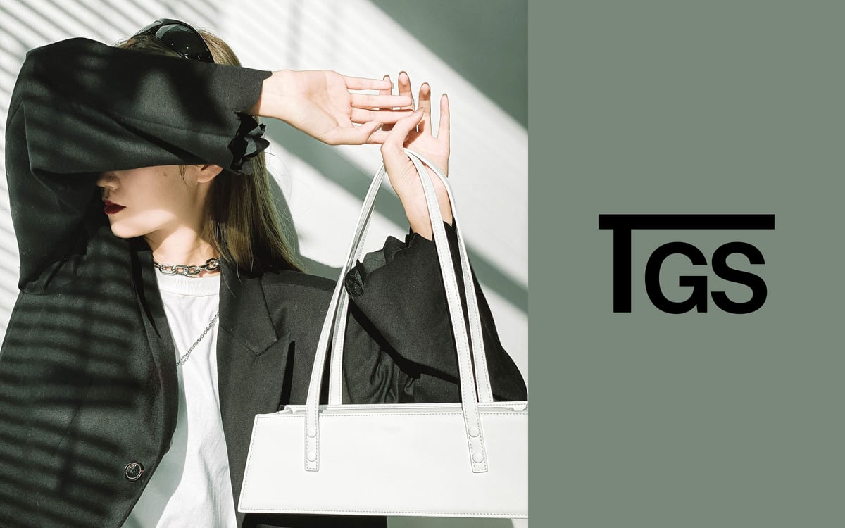 The Good Stuff campaign image: a woman shading her eyes with one arm and holding a white top-handle bag in striped sunlight, beside a sage field carrying the TGS monogram